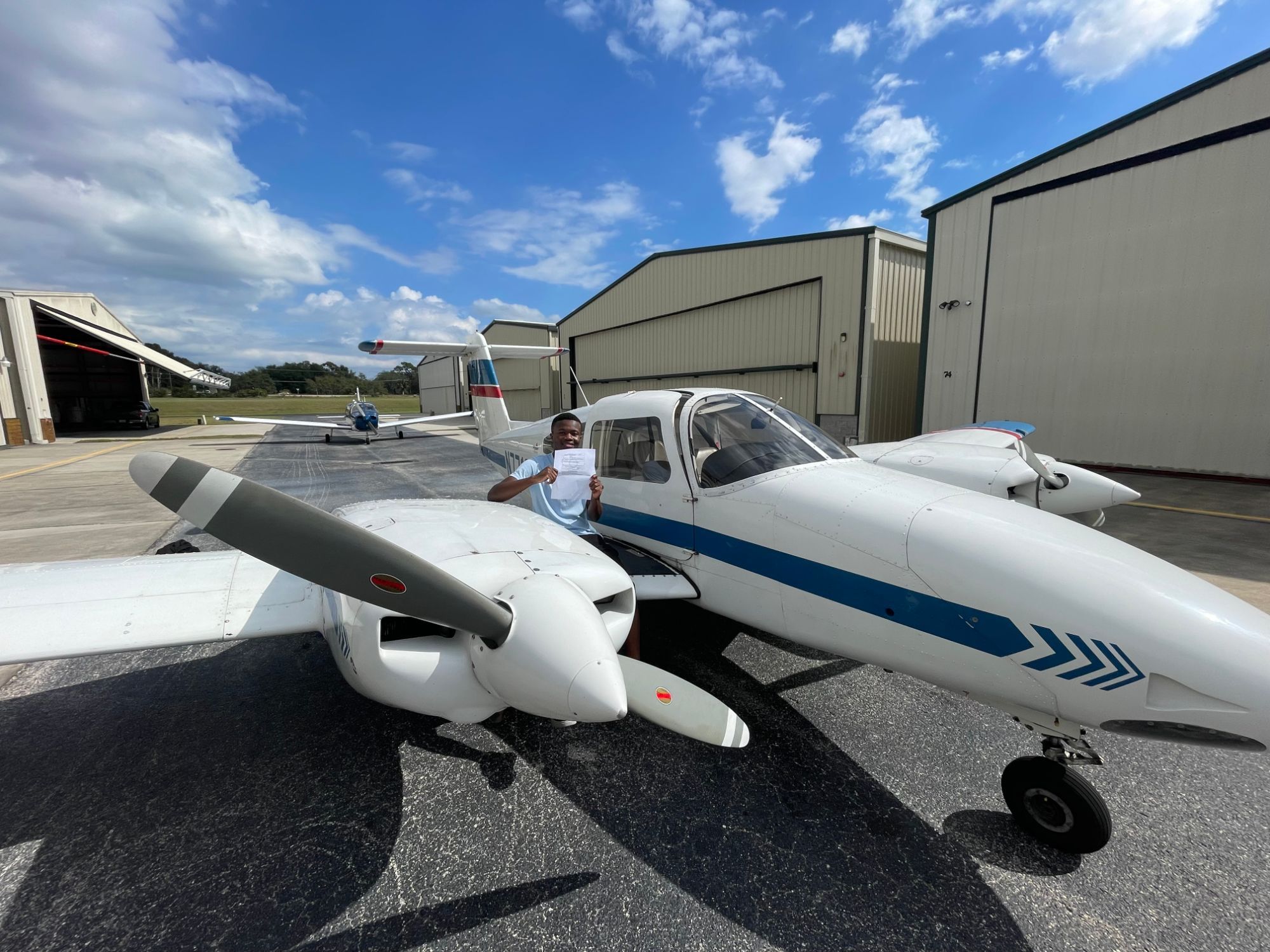 Enoch Kund smiling and holding up a sheet of paper beside a white twin-engine airplane with blue stripes, on a sunny airport ramp lined with hangars.