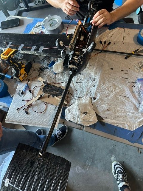 Workbench covered in plastic sheeting; two people work on the opened fuselage, with the carbon tail boom and a carbon tail surface laid out beside it.